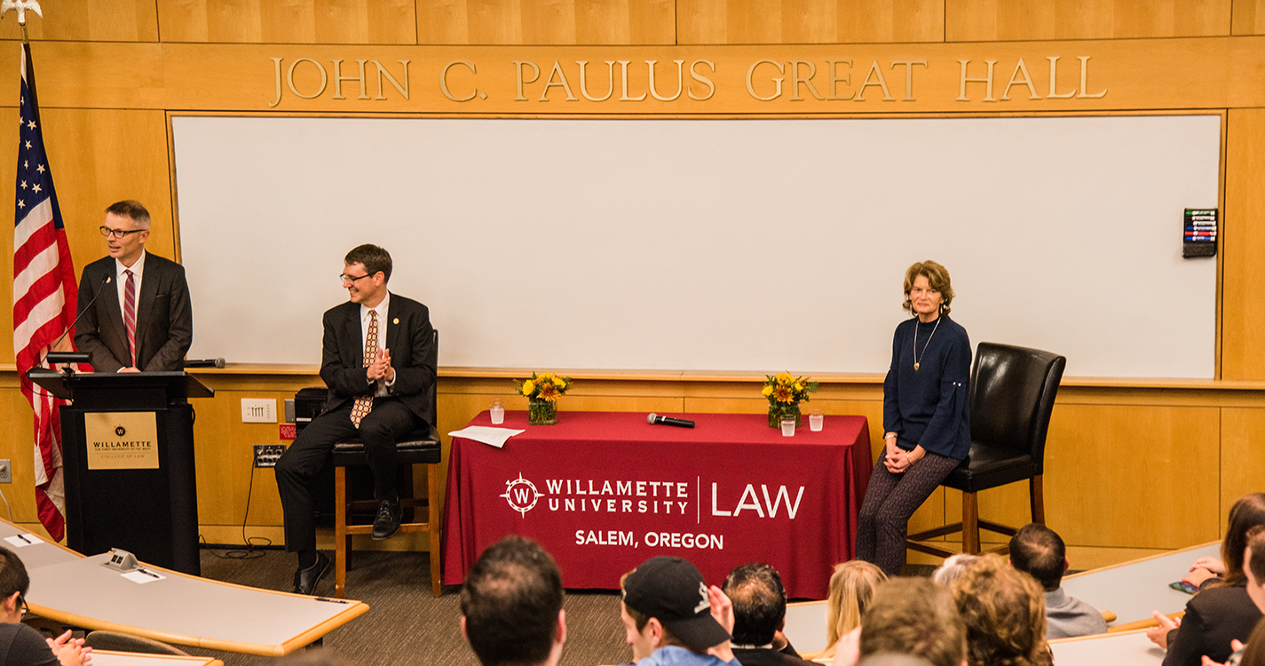 Sen. Lisa Murkowski sits on a chair in front of a lecture hall as Dean Bridgeman introduces her.