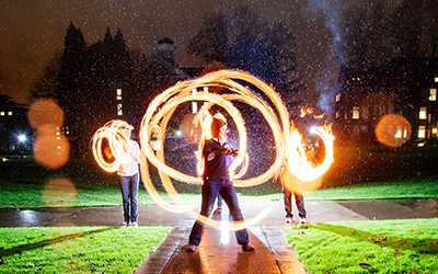 three students twirl flaming poi in a time-lapse photo so the fire forms circles around them