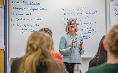 Professor Karen Arabas stands before a classroom with a whiteboard with Overview Colorado River written on it.