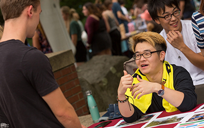 A smiling student seated at a table outside helps a standing student sign up for a club