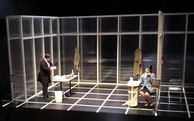 two actors wear masks on a black stage set marked with a white grid