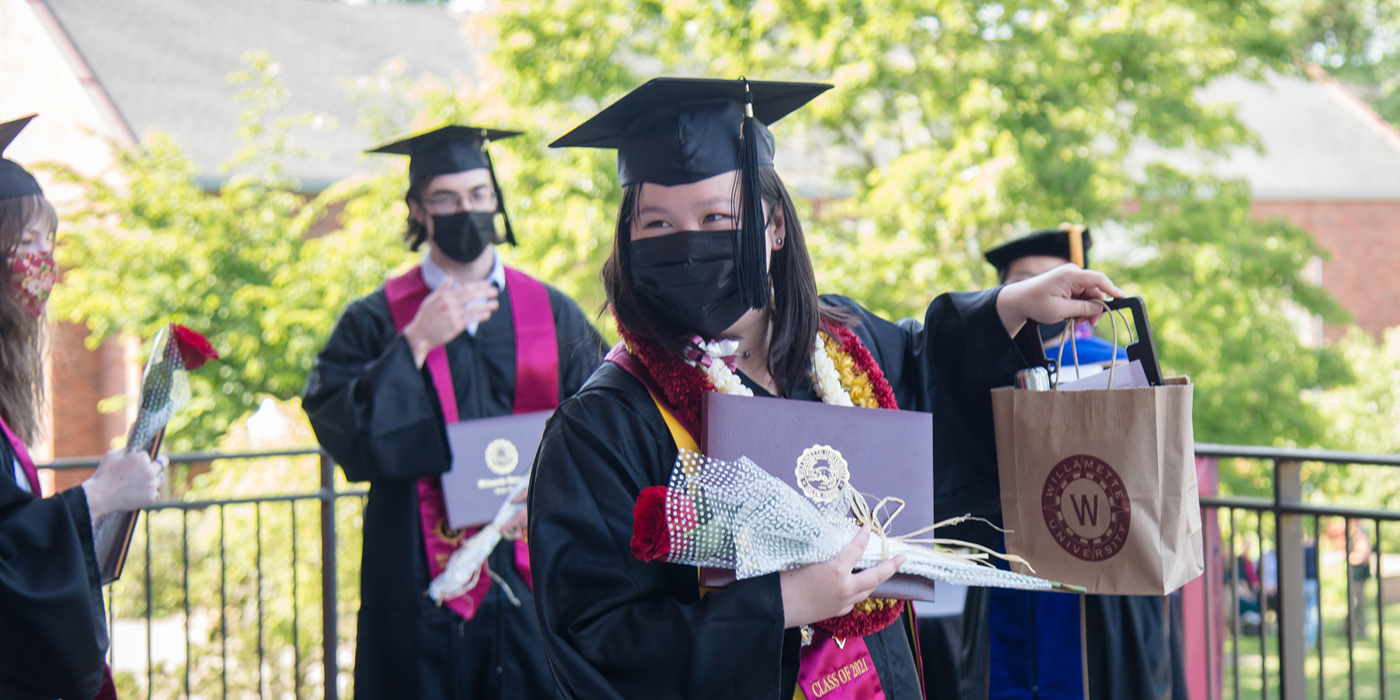 A graduate stands with flowers and a diploma